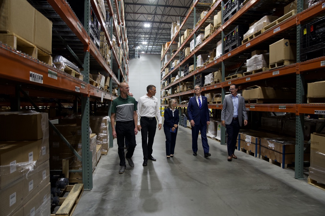 Scott Bessent and a group walking down a wide aisle between tall industrial shelving units filled with boxes and parts.