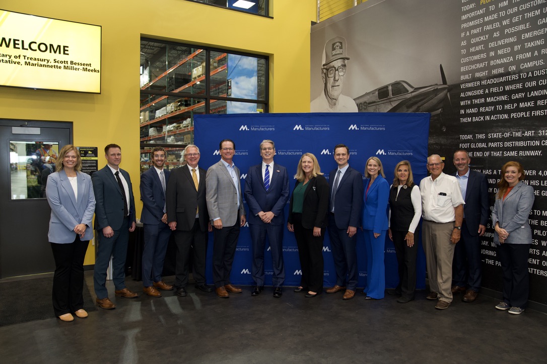 Group photo of Scott Bessent and about a dozen other people posing in front of a blue National Association of Manufacturers backdrop inside a facility.