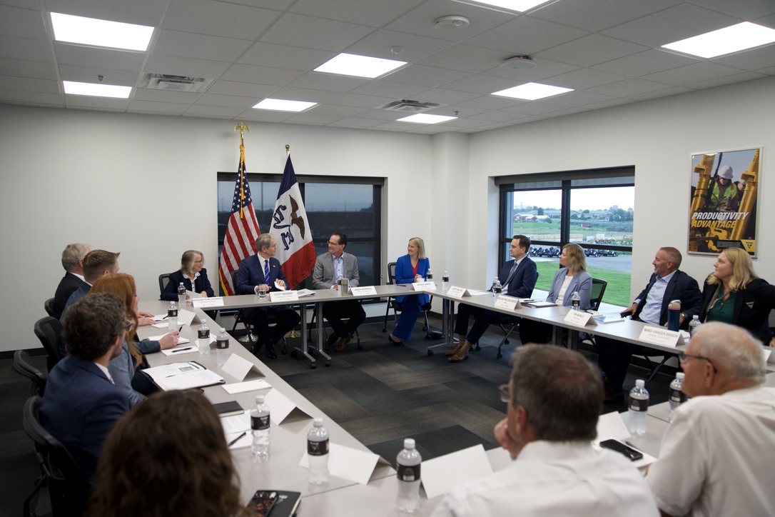 Scott Bessent seated at a conference table in a meeting room with other participants, American and Iowa state flags visible in the background.