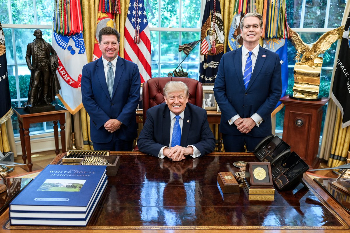 Photo of President Trump seated at the Resolute Desk in the Oval Office, smiling, with Jay Clayton and Scott Bessent standing beside him in suits. American flags, the presidential seal, and other official decor are visible in the background.