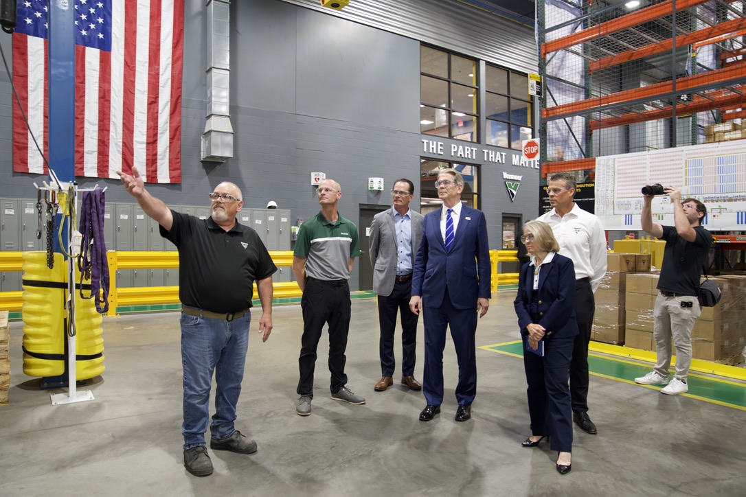 Scott Bessent and a group of people in business and casual attire standing in an industrial facility, with one man pointing towards equipment and an American flag in the background.