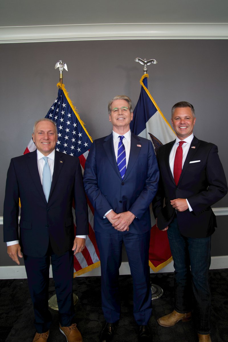 Group photo of three men in suits standing in front of American and Iowa state flags. From left: a man in a dark suit with light blue tie, Scott Bessent in a blue suit with striped tie, and a man in a dark suit with red patterned tie.