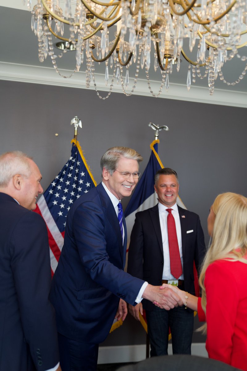 Scott Bessent smiling and shaking hands with a woman in a red jacket, while another man and a man in a red tie look on, with flags in the background.