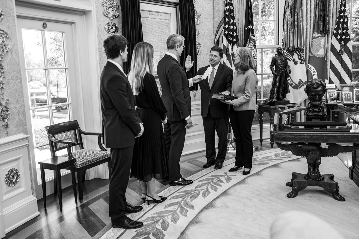 Black and white photo of Jay Clayton being sworn in as Director of National Intelligence in the Oval Office, with his family present, Scott Bessent administering the oath, and official furnishings in the background.