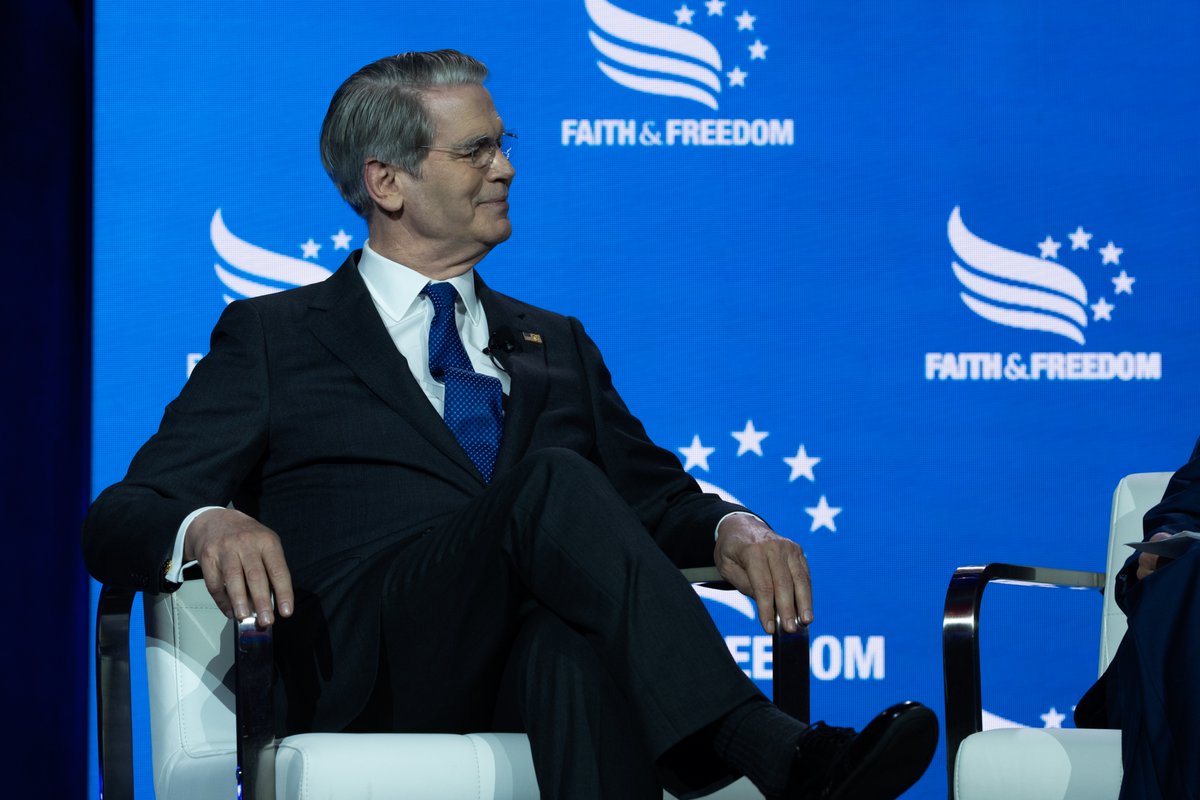 Portrait of Scott Bessent seated in a white chair on stage at the Faith & Freedom event, looking to the side with a slight smile against a branded blue backdrop.
