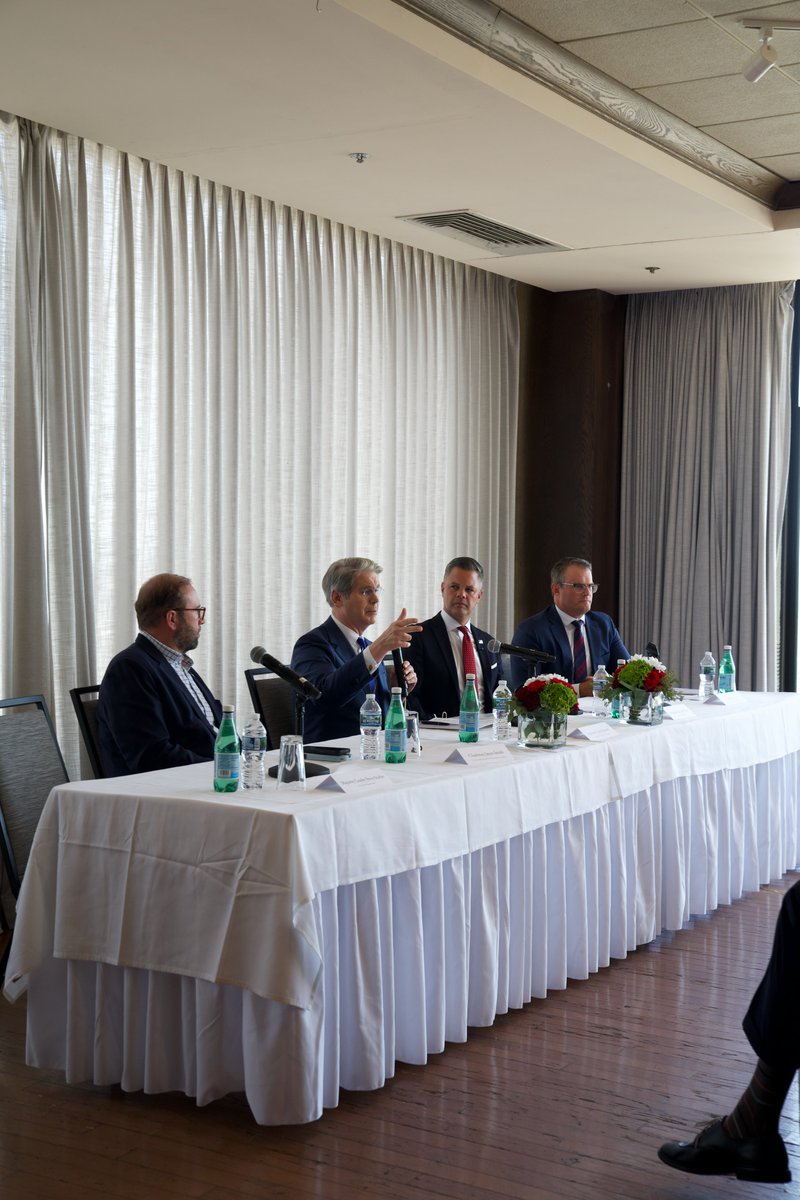 Scott Bessent speaking and gesturing at a long conference table with other panelists, in a room with white curtains.