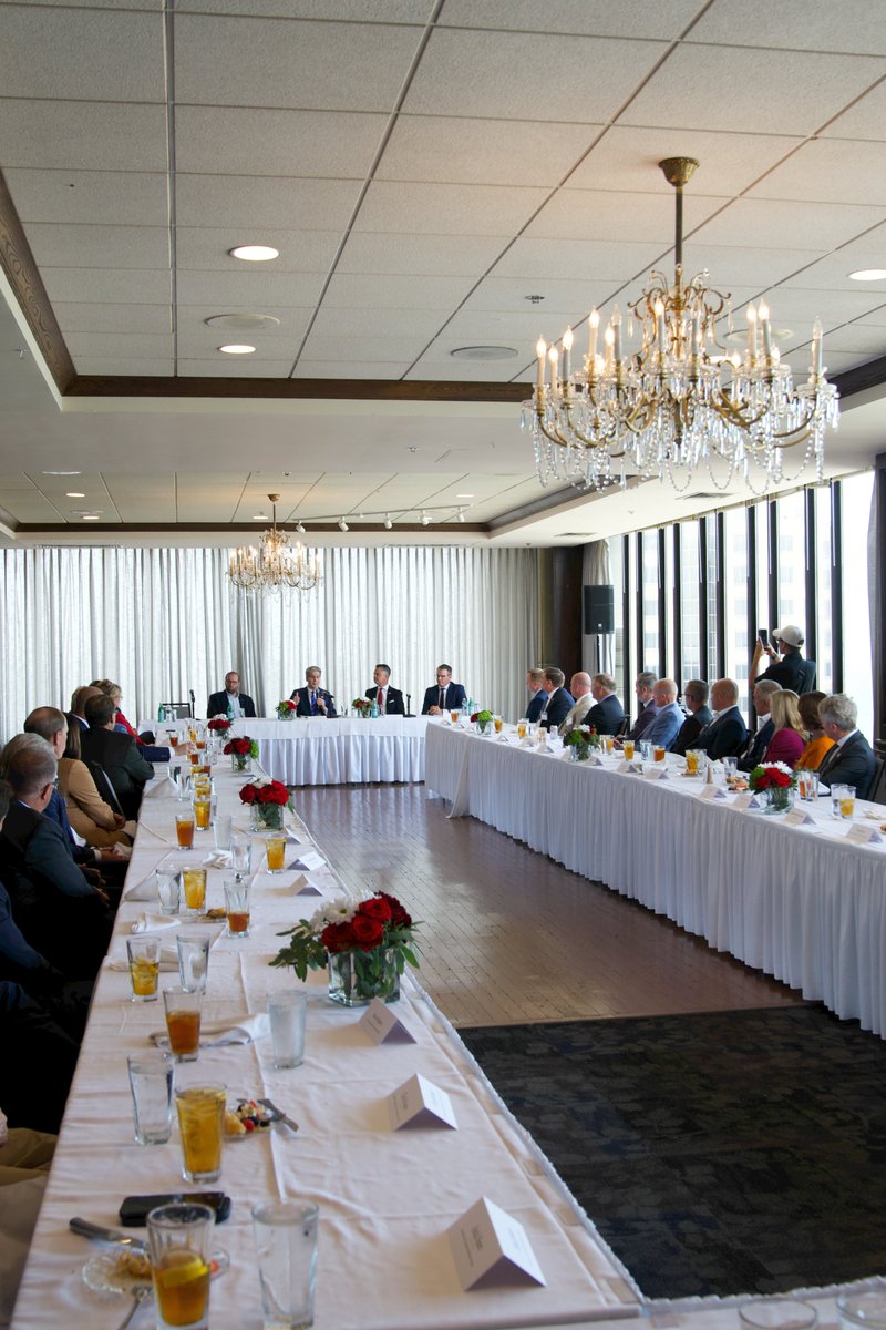 Wide view of a large banquet-style event room with long tables set for dining, chandeliers, and attendees seated.