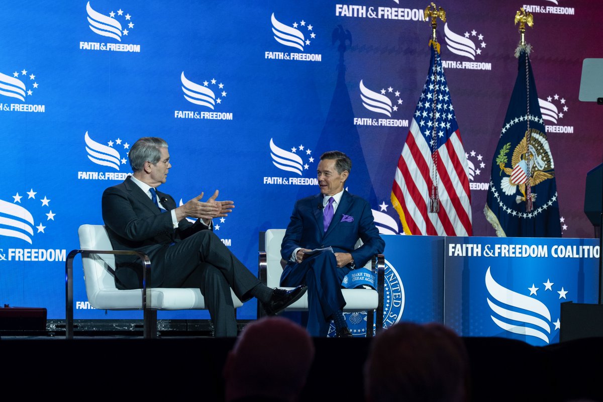 Scott Bessent and Ralph Reed seated on stage at the Faith & Freedom Coalition event, engaged in conversation with American flags and event branding in the background.