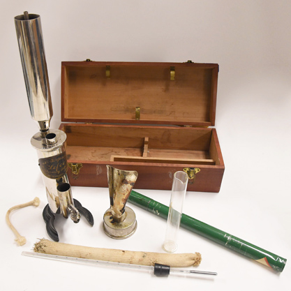 Color photograph of Ebulliometer Device. A wooden box with a metal pull on top and two metal closures which opens to reveal the instrument with parts, and a thermometer in green metal case.