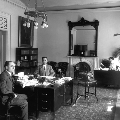 Black and white photograph of 2 men sitting at desk in the Andrew Johnson suite with a typewriter beside them.